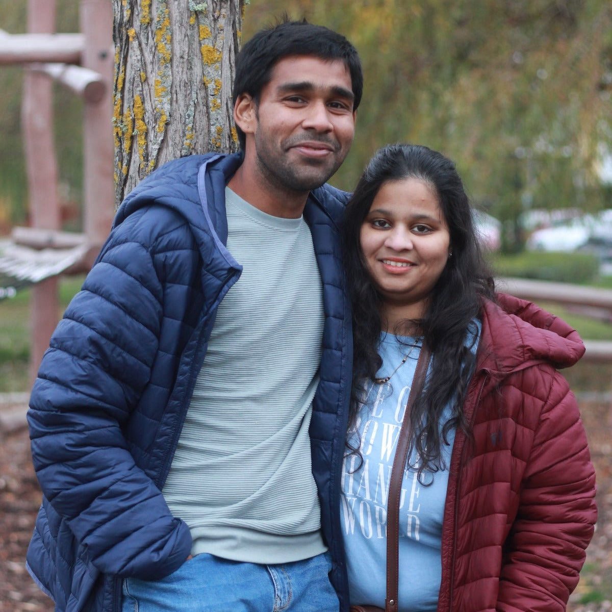 Two people standing close together outdoors, leaning against a tree.